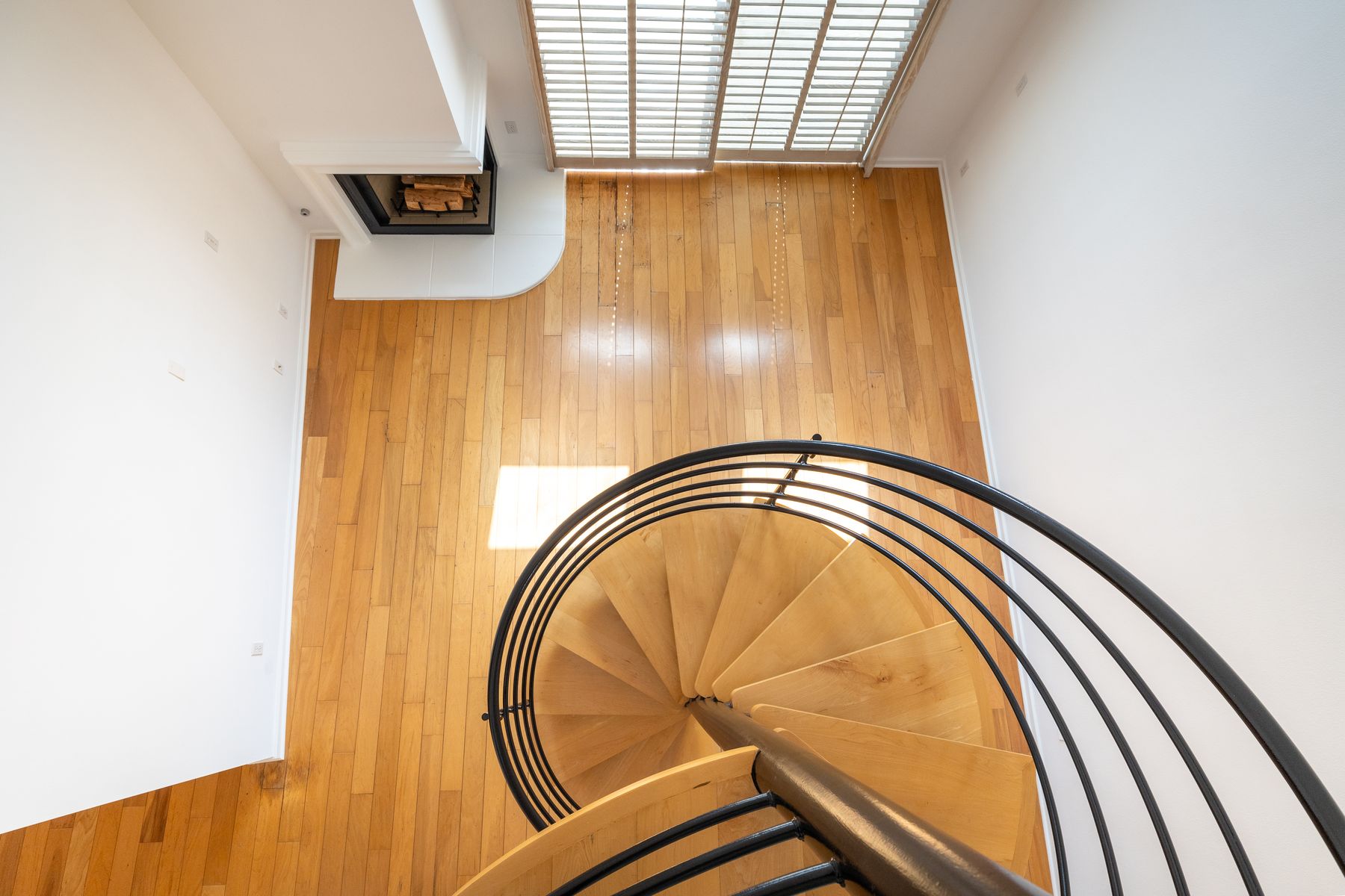 A curved wooden staircase with a dark railing, looking down toward a sunlit wooden floor.